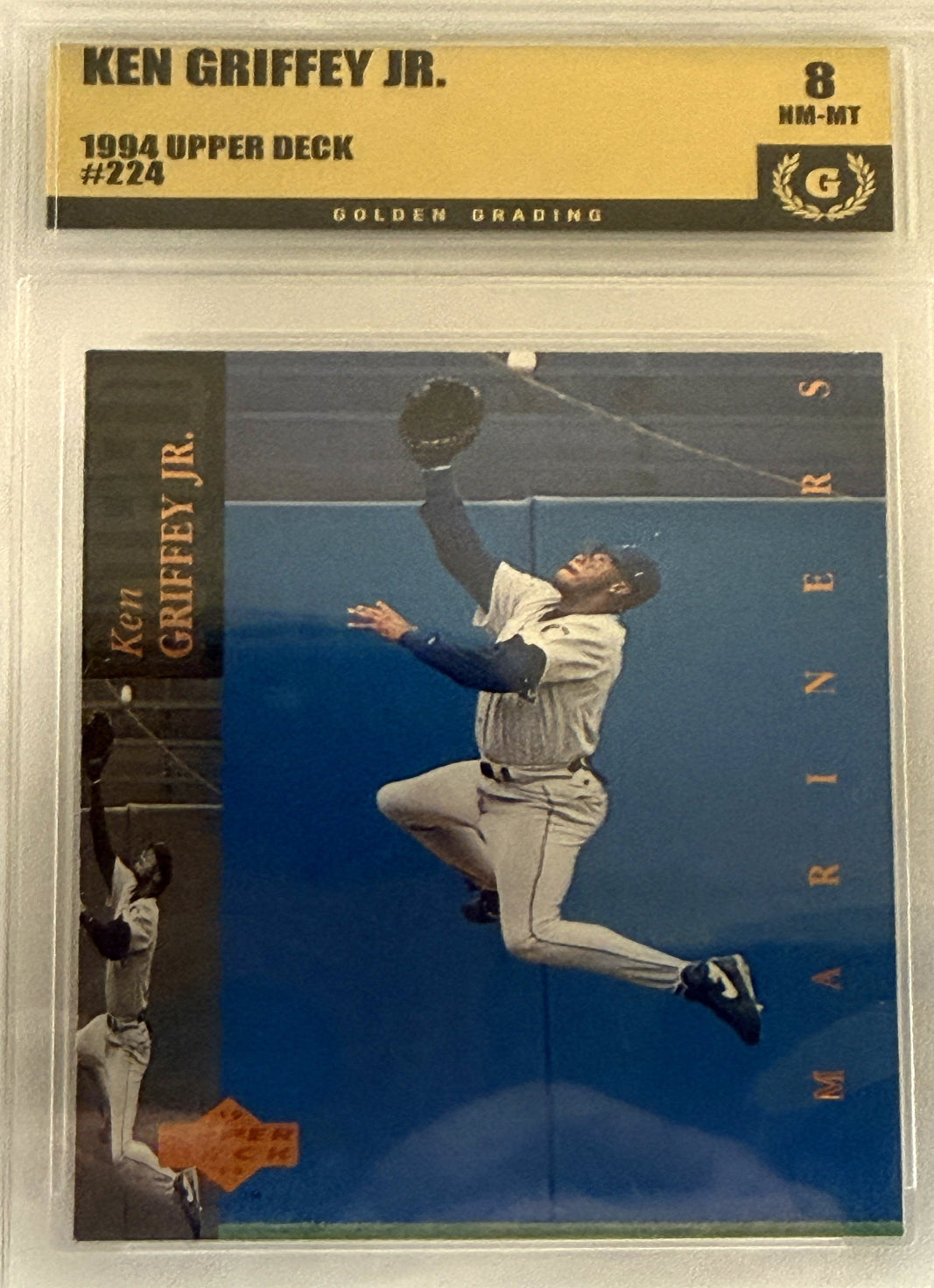 A graded baseball trading card featuring Ken Griffey Jr. in a Seattle Mariners uniform, captured mid-air while reaching for a ball against a blue outfield wall.  Grade 8