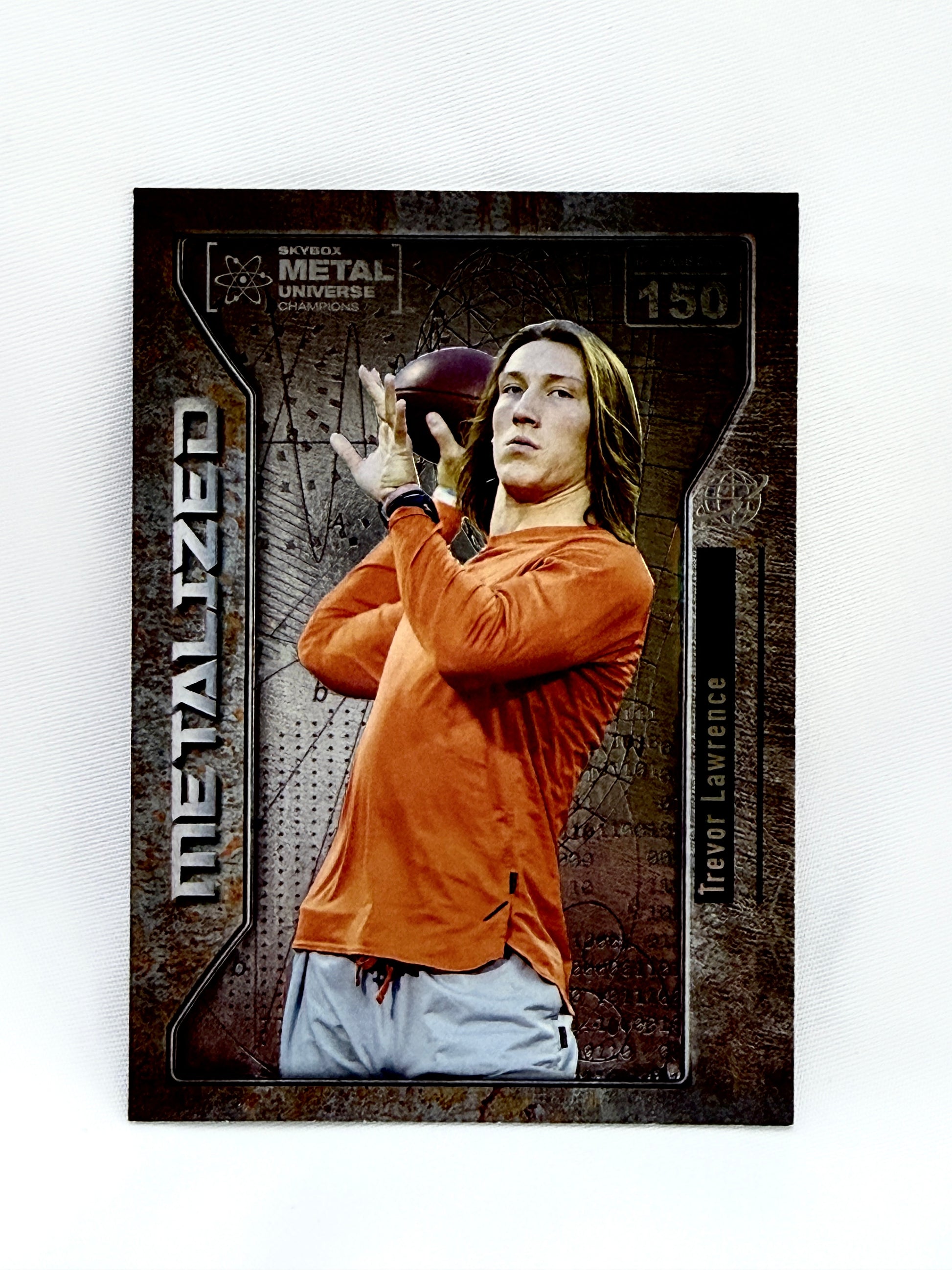 A Trevor Lawrence trading card from the 2012 Metal Universe Draft, featuring a photo of Lawrence in an orange long-sleeve shirt and white pants, set against a dark textured background with 'METALIZED' printed vertically on the left.